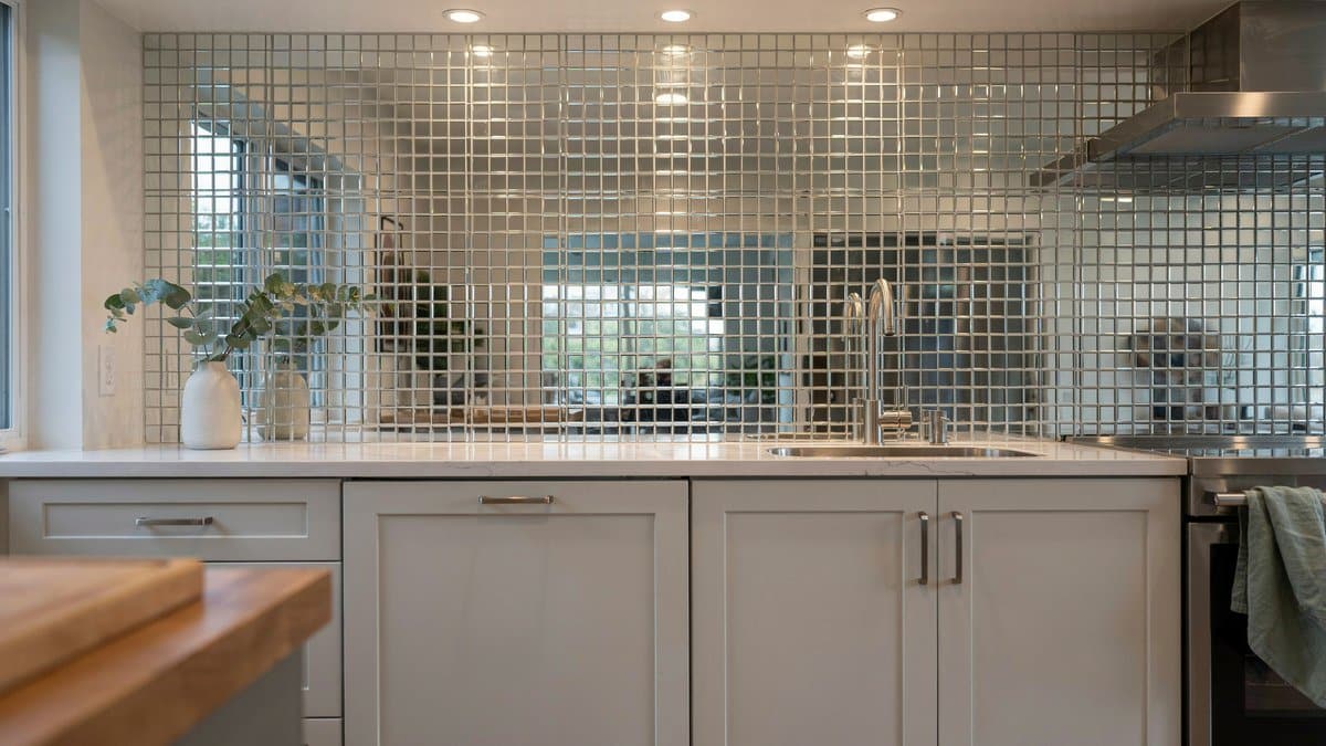 Small mirrored mosaic tile backsplash reflecting a bright white kitchen and recessed lights