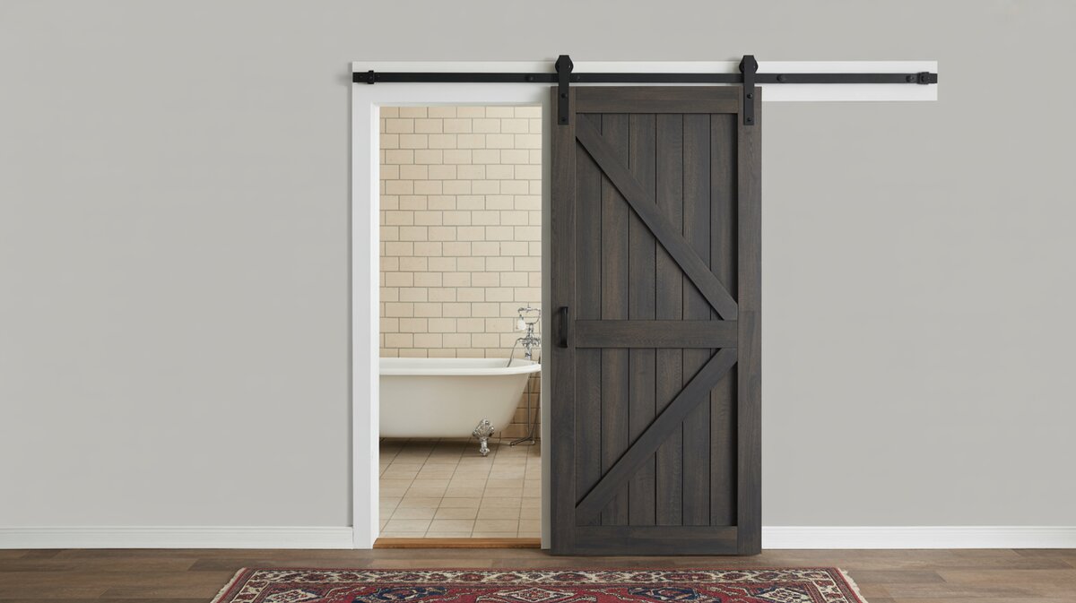 Dark charcoal sliding barn door on black hardware against grey wall partially open to beige tile bathroom