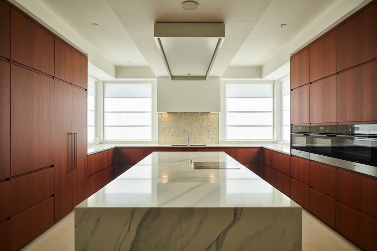 Cherry wood cabinets bracket a veined marble island with a golden mosaic backsplash between windows