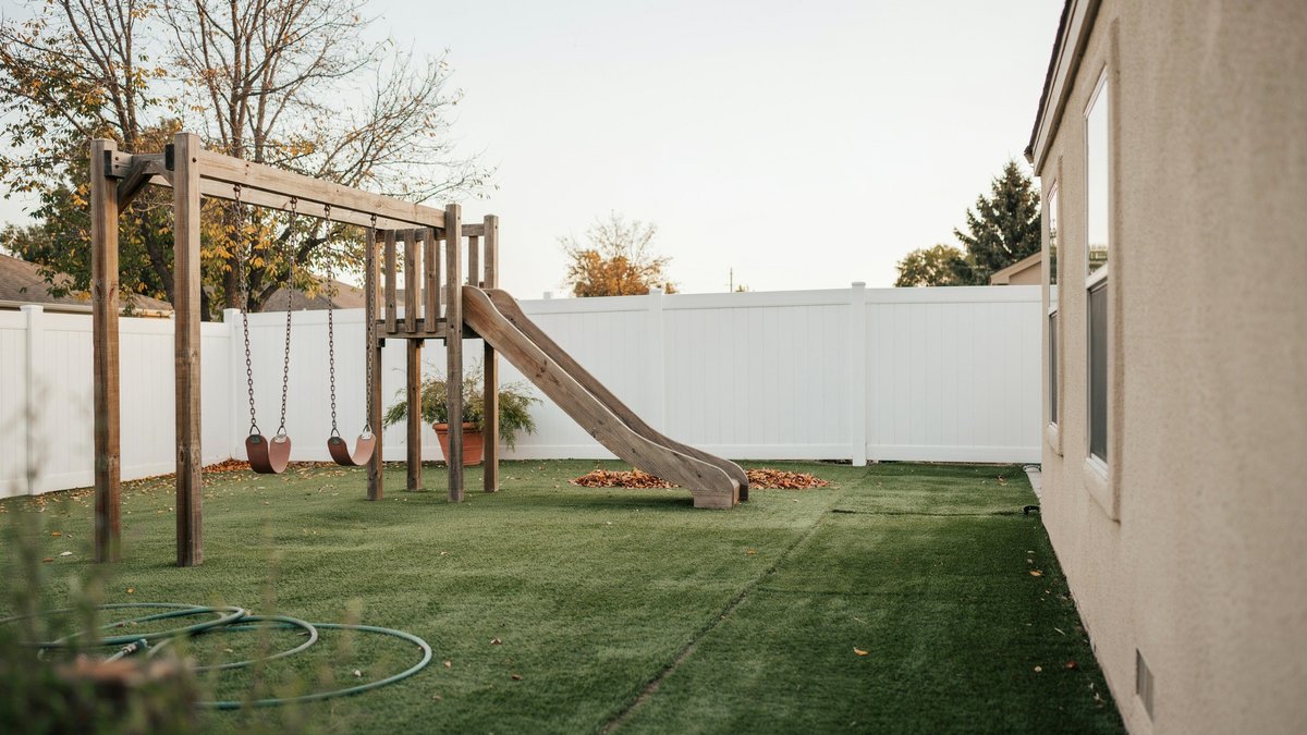 Wooden swing set and slide on turf against a white vinyl fence