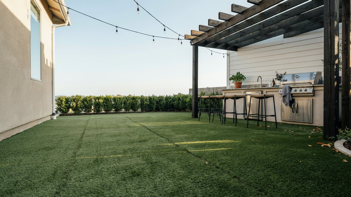 Turf lawn beside a covered outdoor kitchen with grill and bar stools