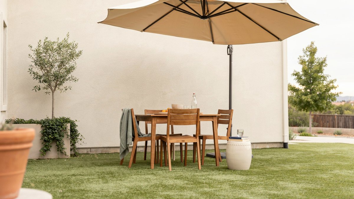 Wood dining set under tan umbrella on turf beside a stucco wall
