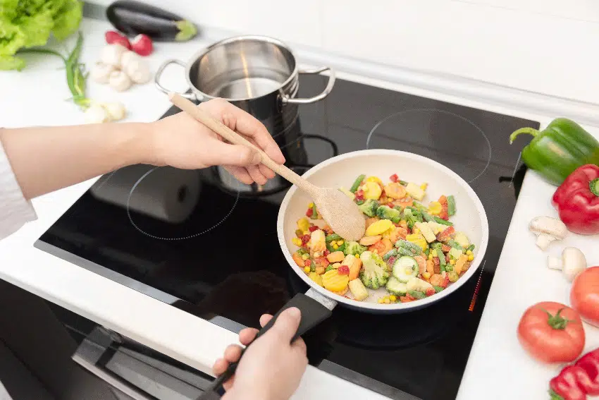 fresh vegetables fried in a pan on a glass ceramic cooktop with some vegetables and herbs around the white kitchen countertop