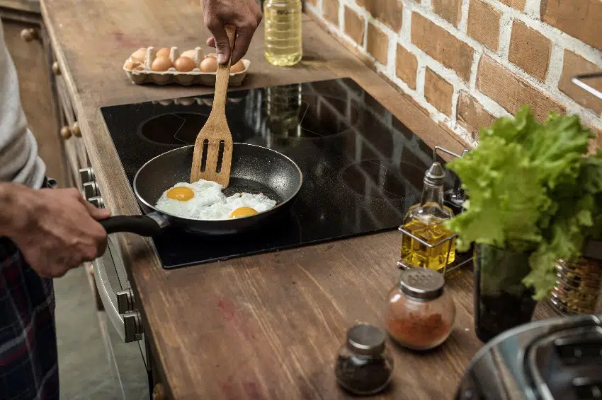 man cooking eggs in a frying pan on a ceramic cooktop for breakfast in wooden kitchen counter