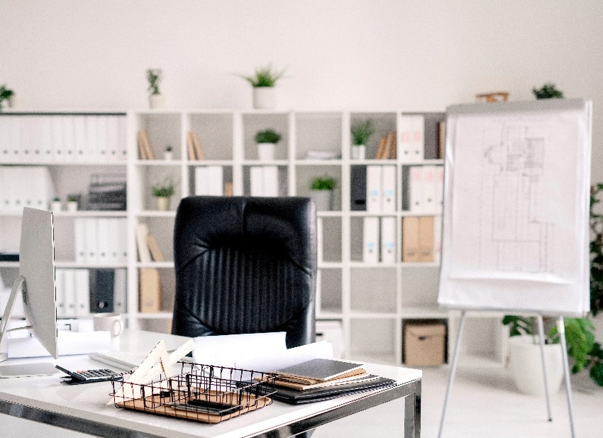 office room with desk, black leather armchair, whiteboard, computer monitor and other supplies on background of shelves with documents