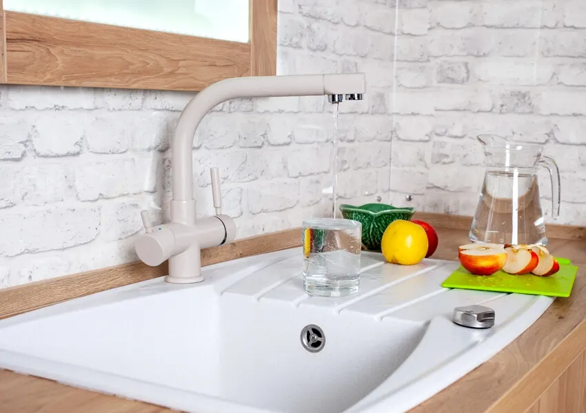 A kitchen with cast iron sink, a faucet with running water, apple slices on chopping board at the side and a glass pitcher with water