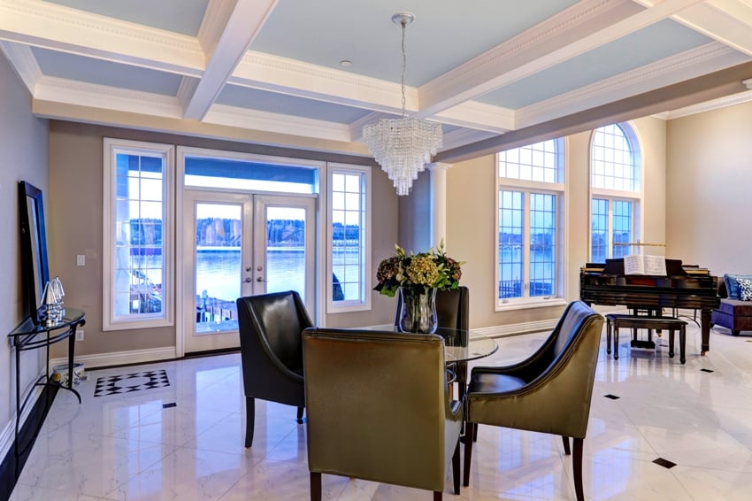 luxury dining area features light blue coffered feature, accented with crystal chandelier illuminating glass top table with leather chairs atop marble floor