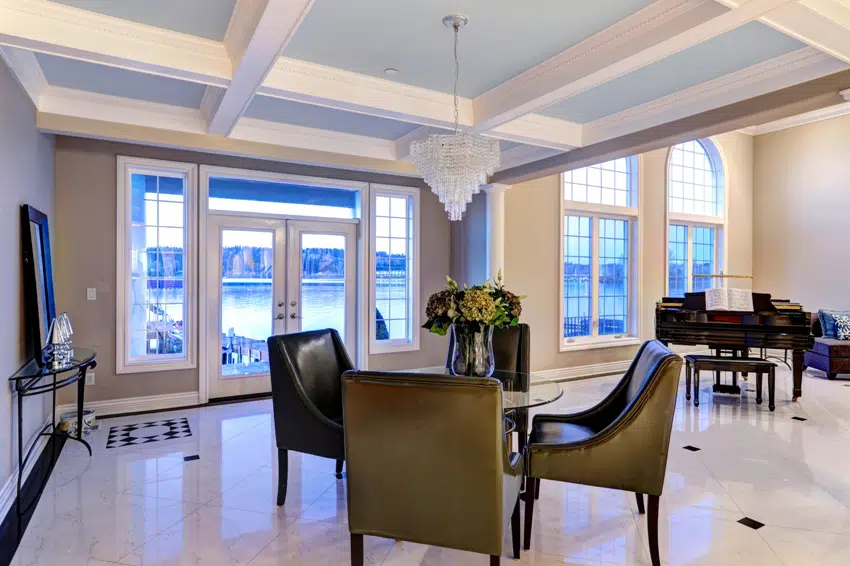 luxury dining area features light blue coffered ceiling accented with crystal chandelier illuminating glass top table with leather chairs atop marble floor