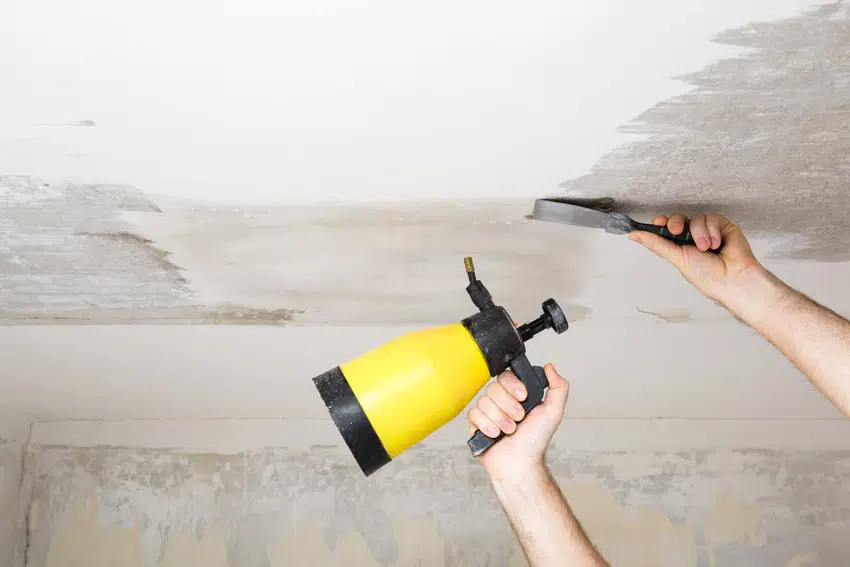 Man removing white chalk residue from ceiling with scraper and sprayer