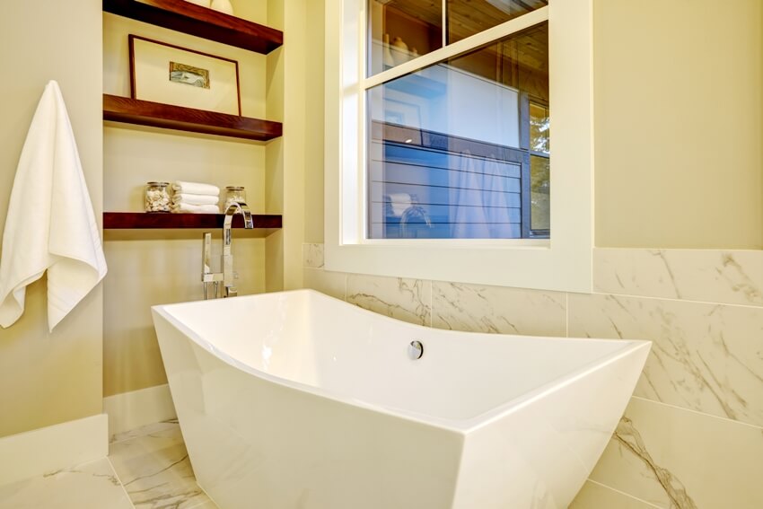 A sophisticated bathroom nook with freestanding tub placed under the window a wall niche with shelves and white and grey marble tiled floor and half wall