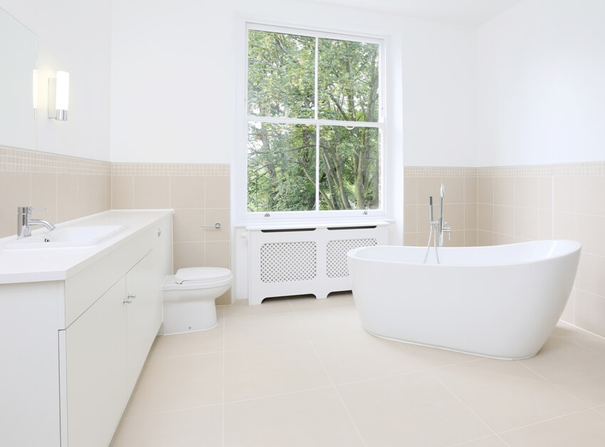 White bathroom with free standing tub, toilet bowl, sink, half tiled walls, and green tree in the window
