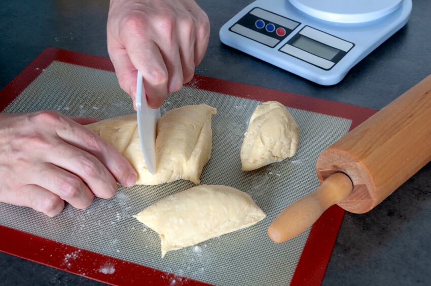 Hands cutting the dough on a silicone mat with rolling pin and weighing scale on the side