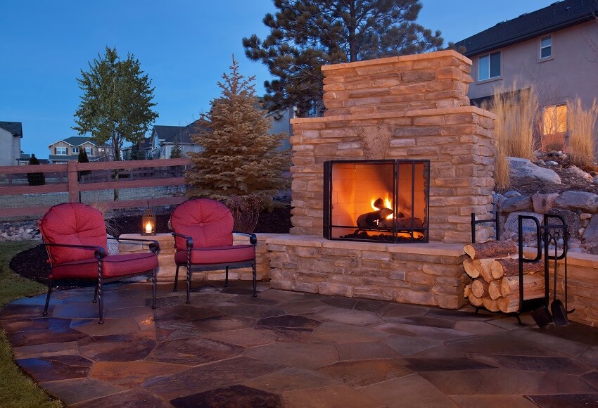 Outdoor stone fireplace covered by a metal grate with two metal chairs with red cushions placed on the flagstone patio