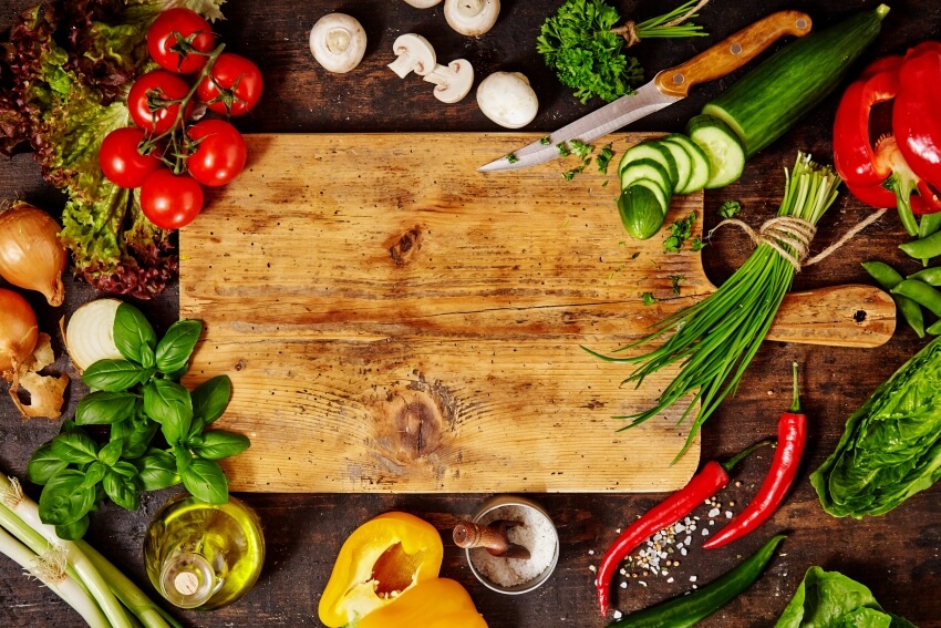 View of knife and wooden cutting board surrounded by fresh herbs and assortment of raw vegetables on rustic wood table