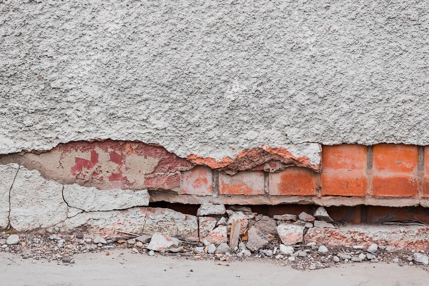 A foundation of a residential building with cracks gradually collapsing and plaster falling off of a brick wall