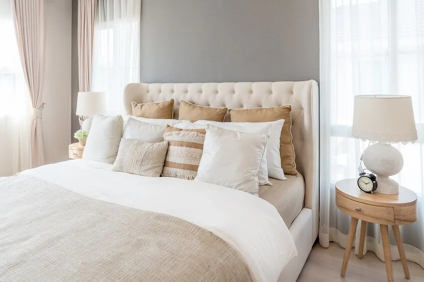Bedroom in soft light colors with lampshade and alarm clock on bedside table and big double bed with pillows and linen sheets