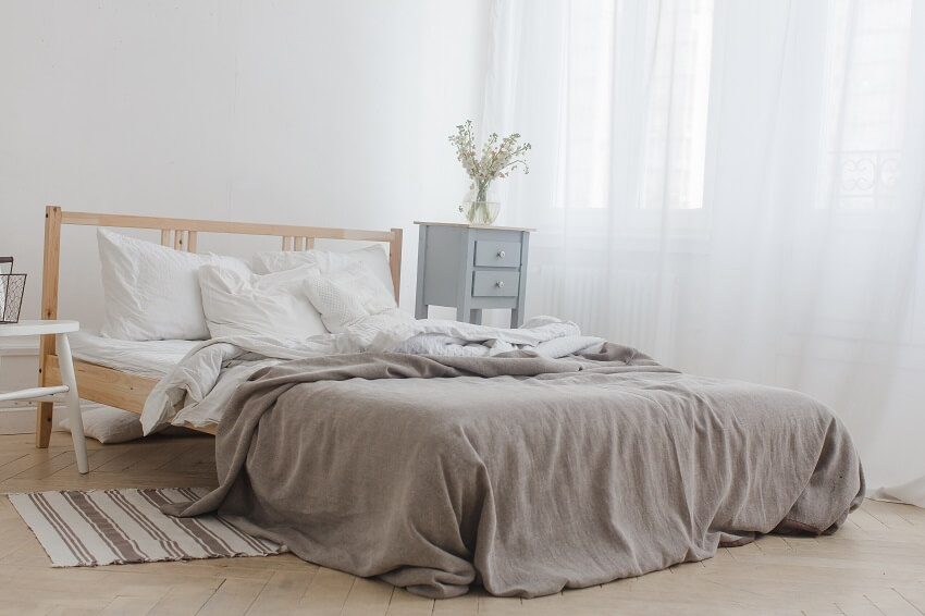 Interior of white and gray cozy bedroom with white curtains wooden bed chair basket flowers bedside table and wooden floor