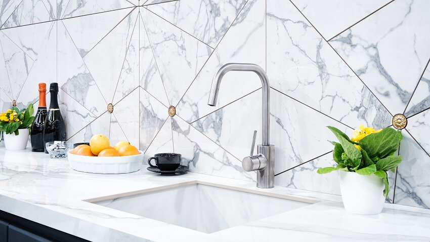 Kitchen with composite marble countertop and backsplash with two bottles of wine white vases with plants a black coffee cup and a bowl of oranges