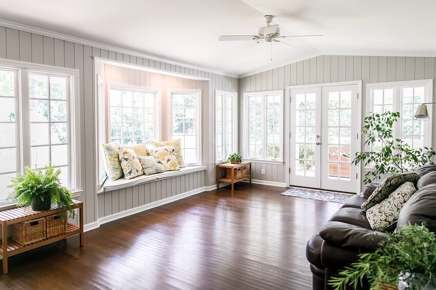 Large and open living room with window seat on one side and a leather couch and plants on the other and a gray panel wall