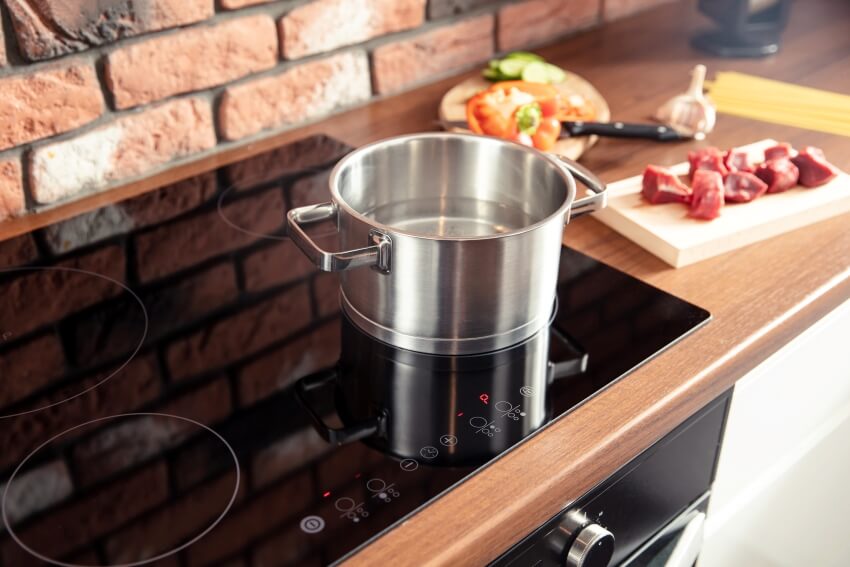 Metal pot stands on a modern induction cooker with meat and veggies on th side