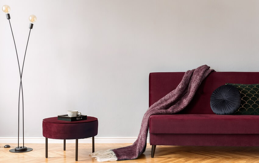 Minimalistic home interior with grey wall maroon sofa ottoman black lamp blanket pillows and brown wooden parquet floor