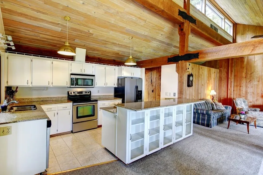 Open kitchen with post and beam tile flooring white cabinets stainless steel appliances pendant lights and view of the living room in a log cabin