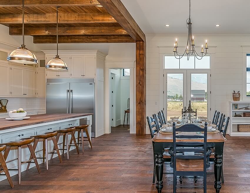Open kitchen with post and beam wood flooring long island with wooden countertop bar stools dining table lighting fixtures and glass double door