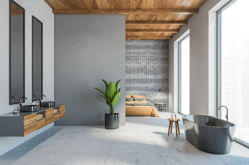 Open space loft apartment with grey living room grey bathtub two sinks brown bed against brick wall and brown wooden ceiling