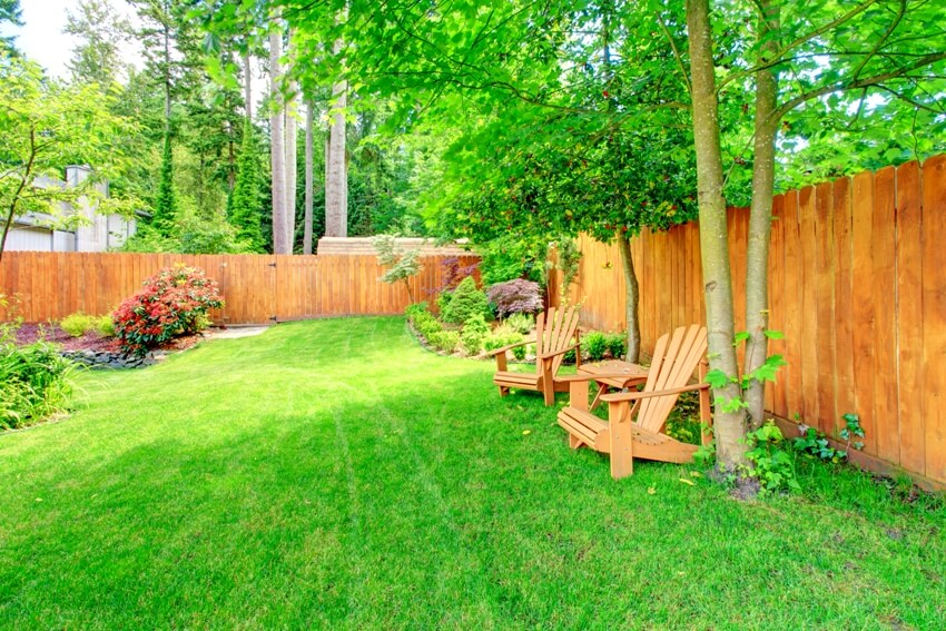 Shiplap fenced backyard with green lawn and flower beds with wooden chairs and table area