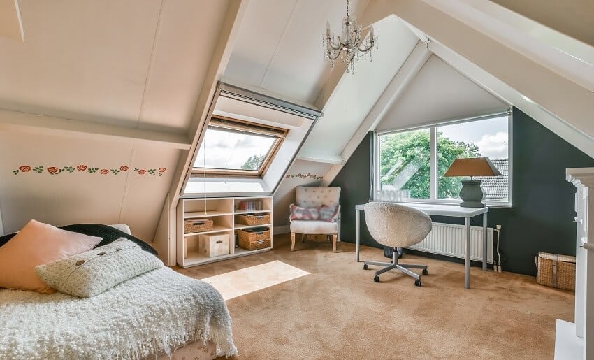 A cozy attic bedroom with carpet lighting fixtures bed shelves table with chair and an armchair by the window