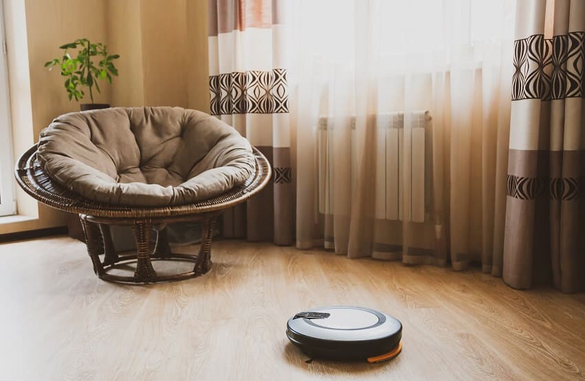 An apartment room with a robot vacuum cleaner on laminate floor curtains and a papasan chair