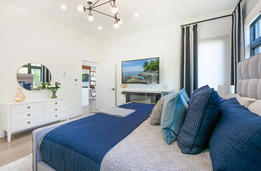Bedroom with blue and white tones a console with vase and flowers on top round mirror lighting fixtures a tv and a cozy large bed