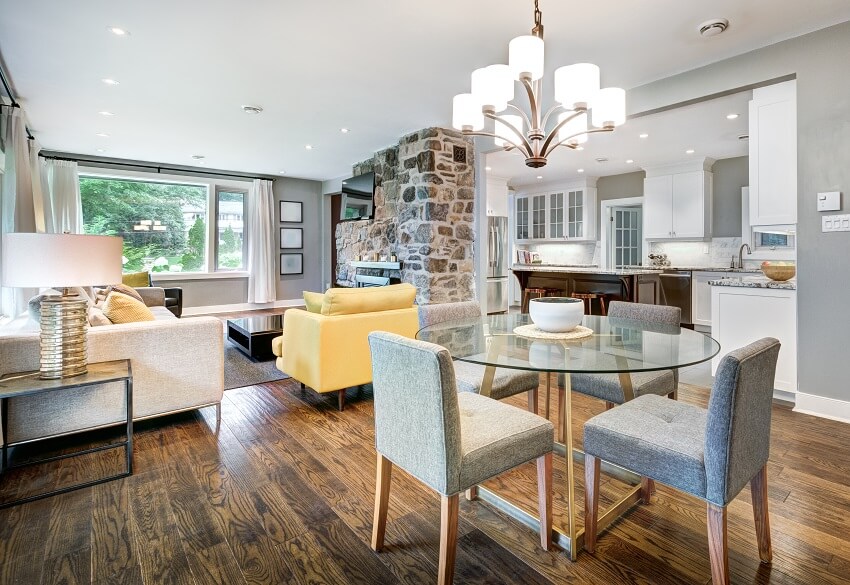 Dining area with a round glass table and chairs chandelier hanging above wood floors grey walls and view of the living room with a stone fireplace