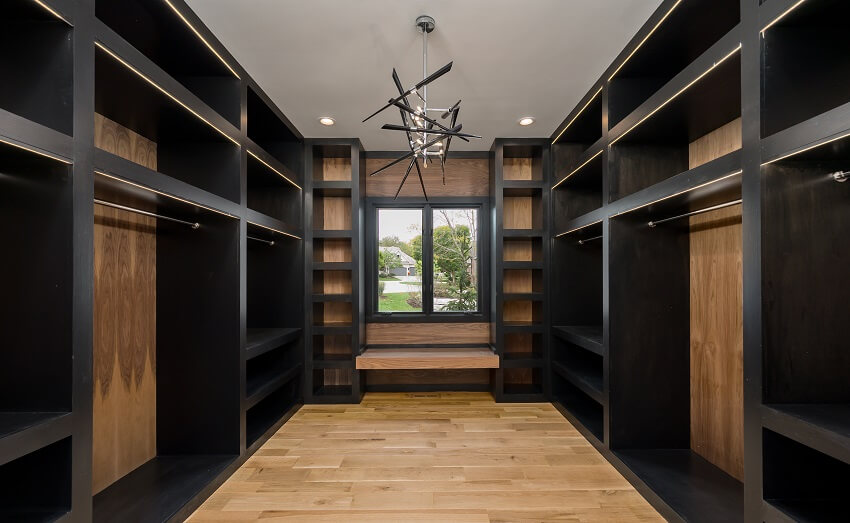 Empty wooden closet with light wood floors bench window lighting fixtures and dark wood shelves closet dividers with metal rod