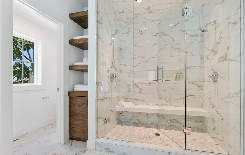 Full glass entry to shower with a long white bench two showerheads marble tile walls and mosaic tile floors