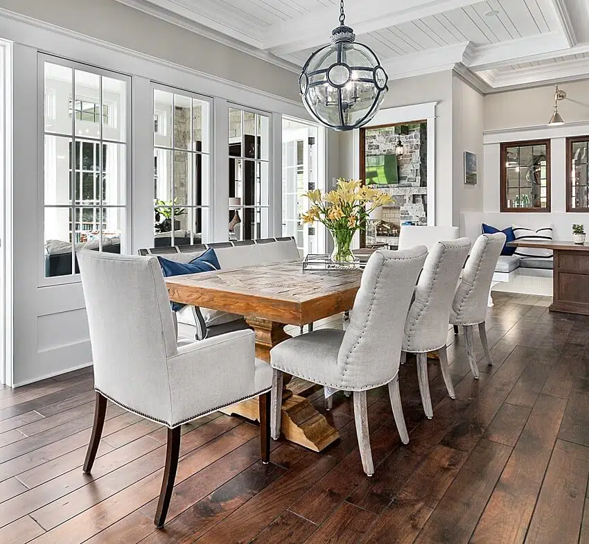 Hardwood floors coffered ceiling and wood dining table with different styles of chairs in a dining and spacious area next to a sunroom