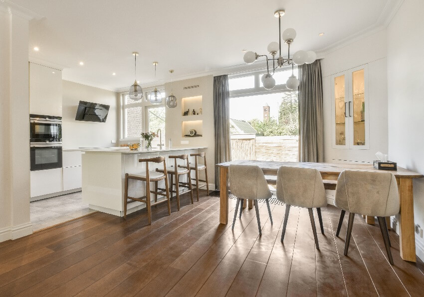 Kitchen and dining area with island wood dining table bar stools large glass windows lighting fixtures white wall and hardwood floors