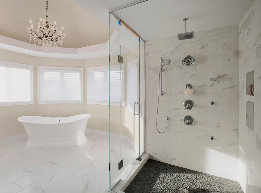 Large master bathroom in a residential home with two shower heads enclosed in a glass with stone floor marble tile wall and a free standing tub with a chandelier above