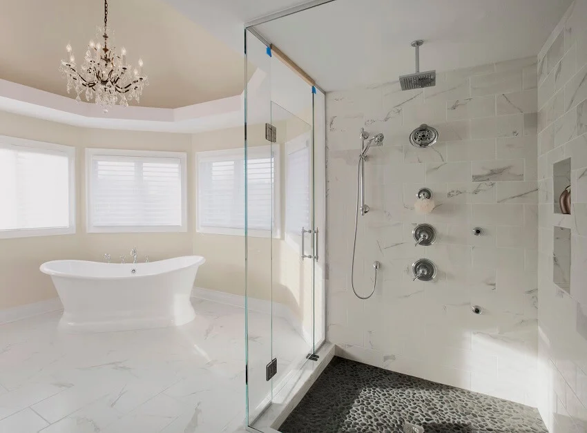 Large master bathroom in a residential home with two shower heads enclosed in a glass with stone floor marble tile wall and a free standing tub with a chandelier above