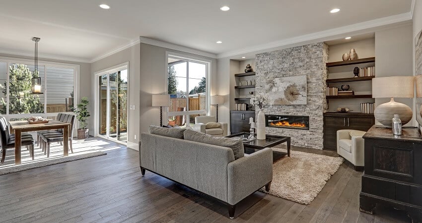 Living room interior in gray and brown colors features gray sofa atop dark hardwood floors facing stone fireplace with built in shelves and view of the dining area