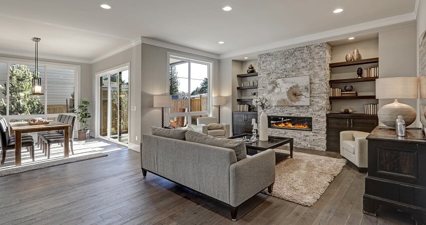 Interior in gray and brown colors features gray sofa atop dark floors facing stone fireplace with built in shelves 