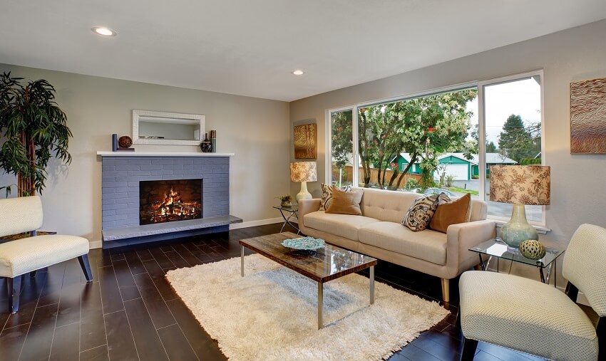 Living room with brick fireplace beige sofa coffee table fur carpet indoor plant view of the outside through large glass window and hardwood floors