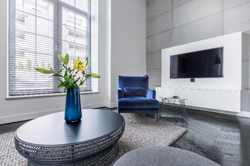 Living room with concrete walls dark wood floors blue armchair blue vase with flower on coffee table and window with blinds