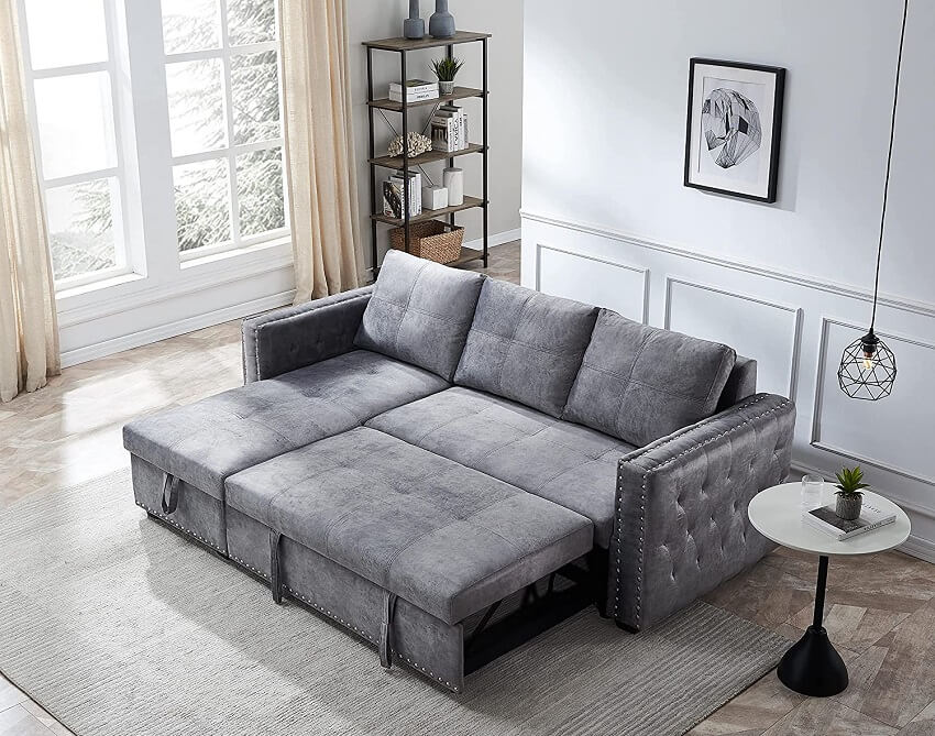 Room with grey velvet L shape reversible sleeper sofa, white table, pendant light, shelves, grey carpet and a large window