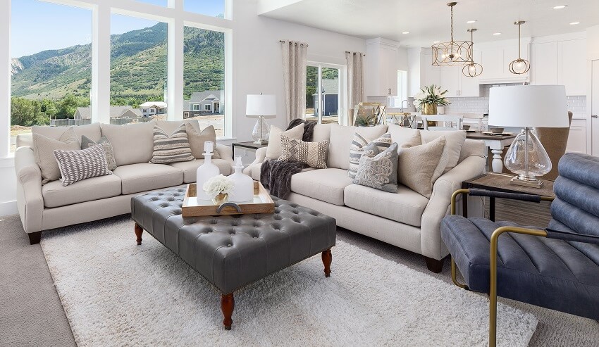 Open concept room with high ceiling, white walls, blue armchair, foot stool ottoman, sofa with cushions, lighting fixtures and a mountain view through large windows