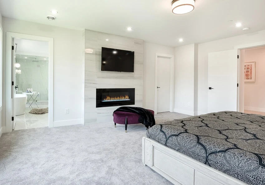 Luxurious carpeted white bedroom with fireplace and tv in a marble wall plum ottoman and view of the bathroom