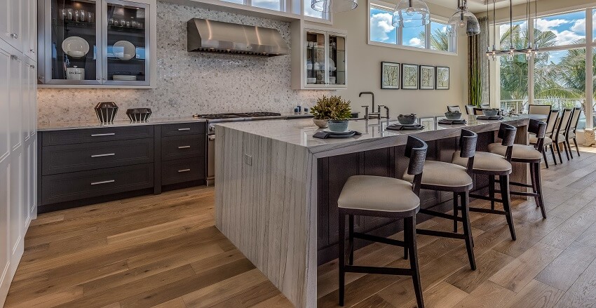 Luxurious kitchen with a large waterfall quartz countertop island barstools wood floors pendant lights and mosaic tile backsplash