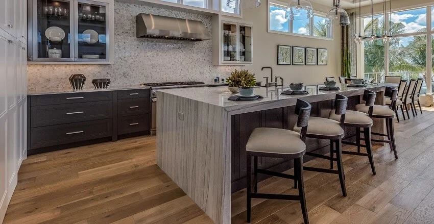 Luxurious kitchen with a large waterfall quartz countertop island barstools wood floors pendant lights and mosaic tile backsplash
