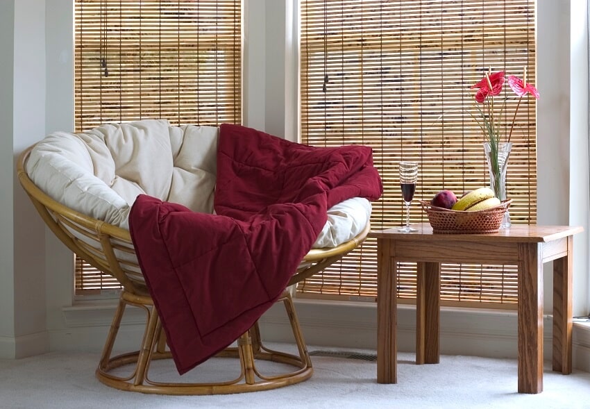 Papasan chair with red throw blanket near coffee table with wine fruits and flower on top in a window with brown blinds background