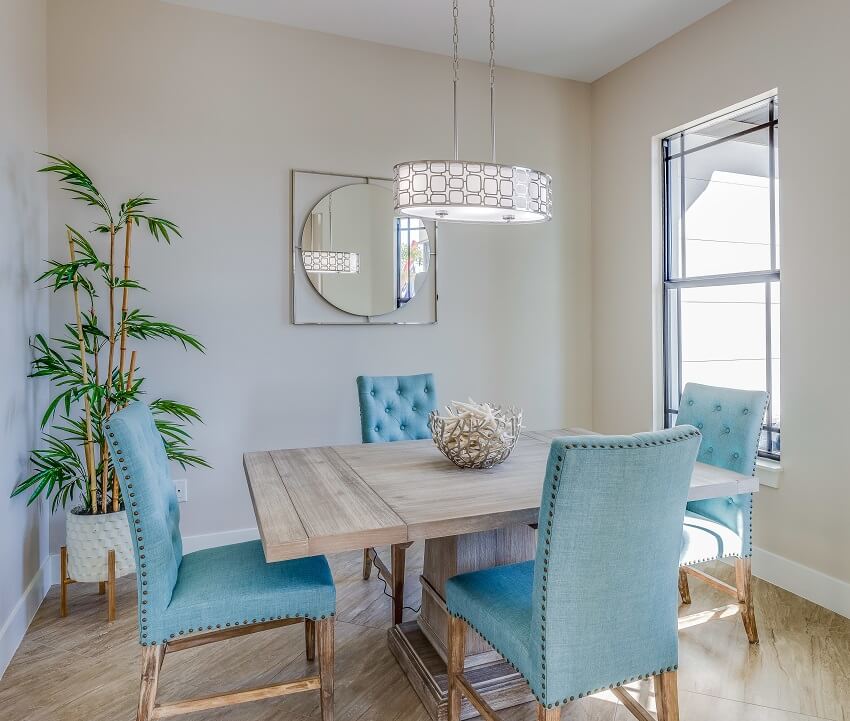 Small and elegant dining room with a square wood table with light blue chairs wood floors pendant light mirror on the wall and an indoor plant